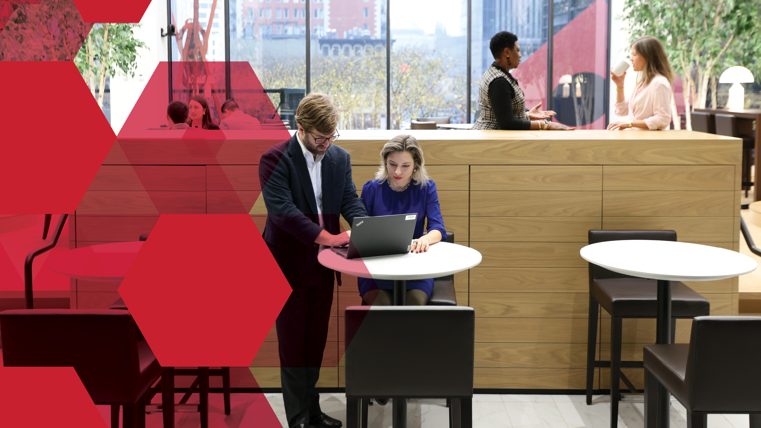 Two people are collaborating at a small round table with a laptop in a modern office. There are others in the background conversing.