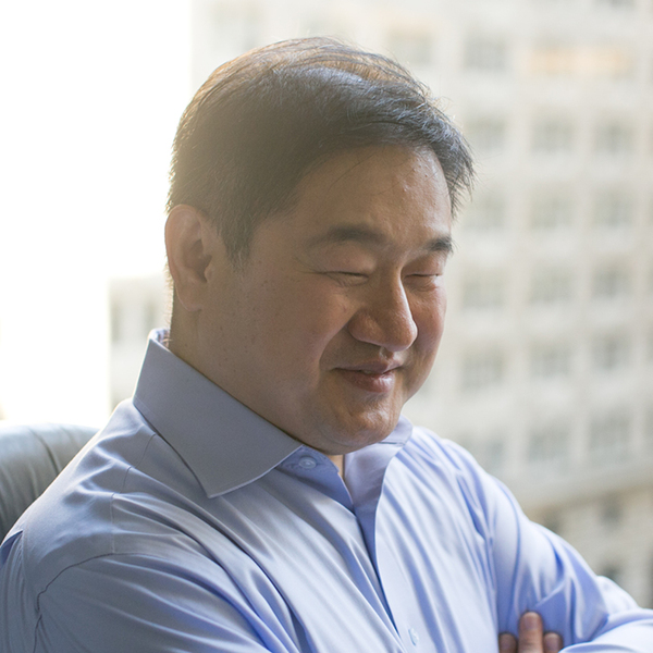 SK Shin with his arms folded, sat in an office chair. There's a view of skyscrapers outside his office window