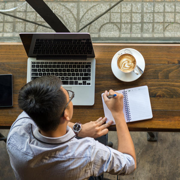 Young Asian worker taking notes beside laptop on wooden table; Remote work concept