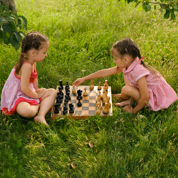 Two girls are playing chess in the garden on the grass. An old wooden chessboard with pieces. Children's games on vacation. The rules of the game. Logical thinking. Think through the moves of the game