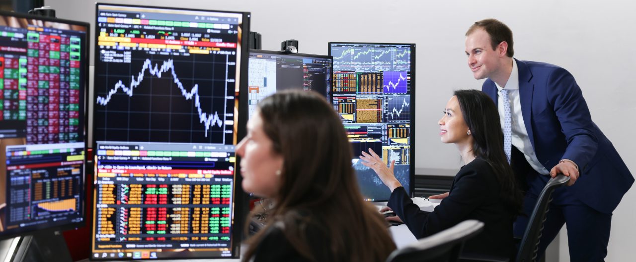 One women in foreground looking at computer screens and a man and a woman in foreground looking at computer screens of charts
