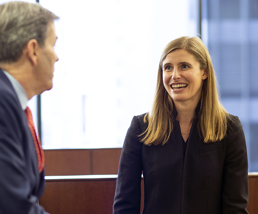 Male and female employee, in business suits, standing at work station desk having a conversation.