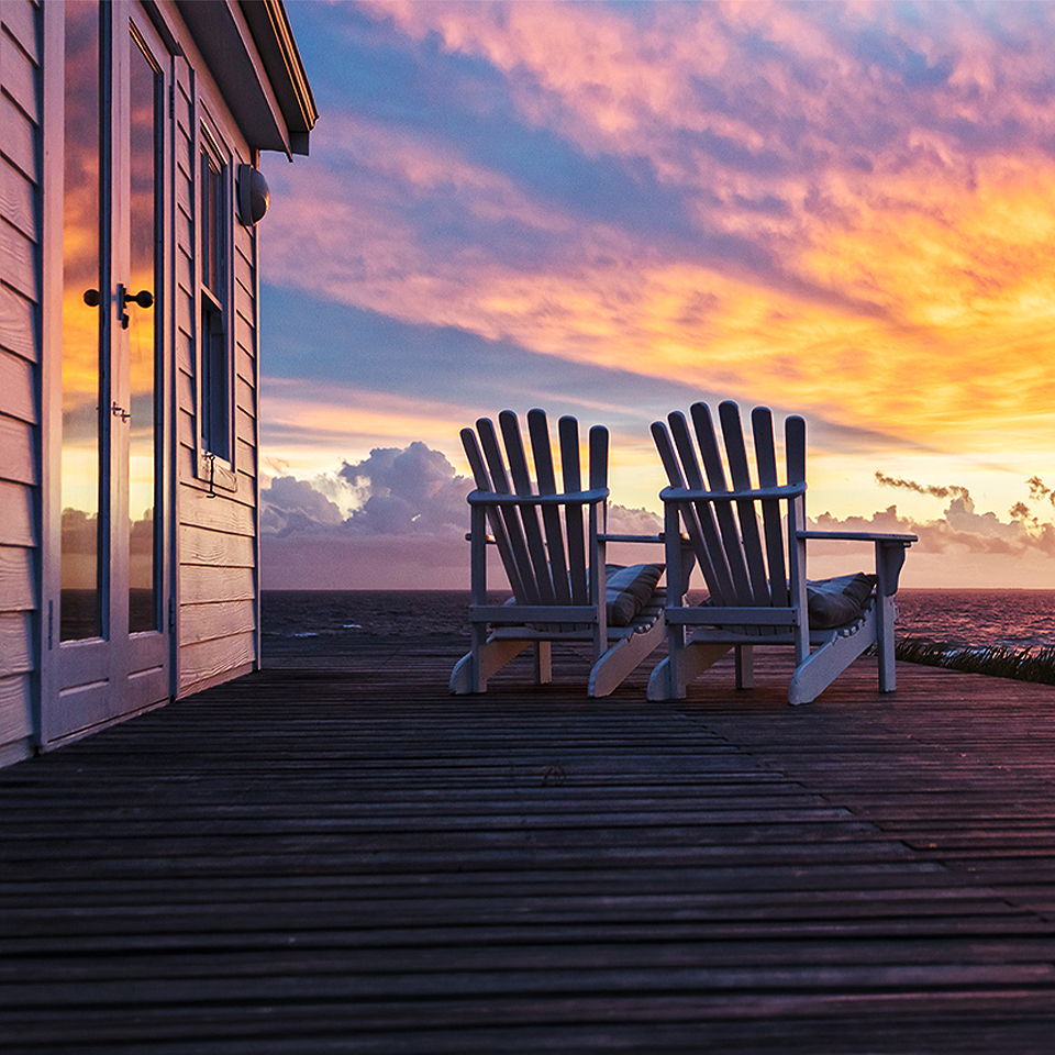 Two Adirondack chairs next to a house on the water and the sun setting